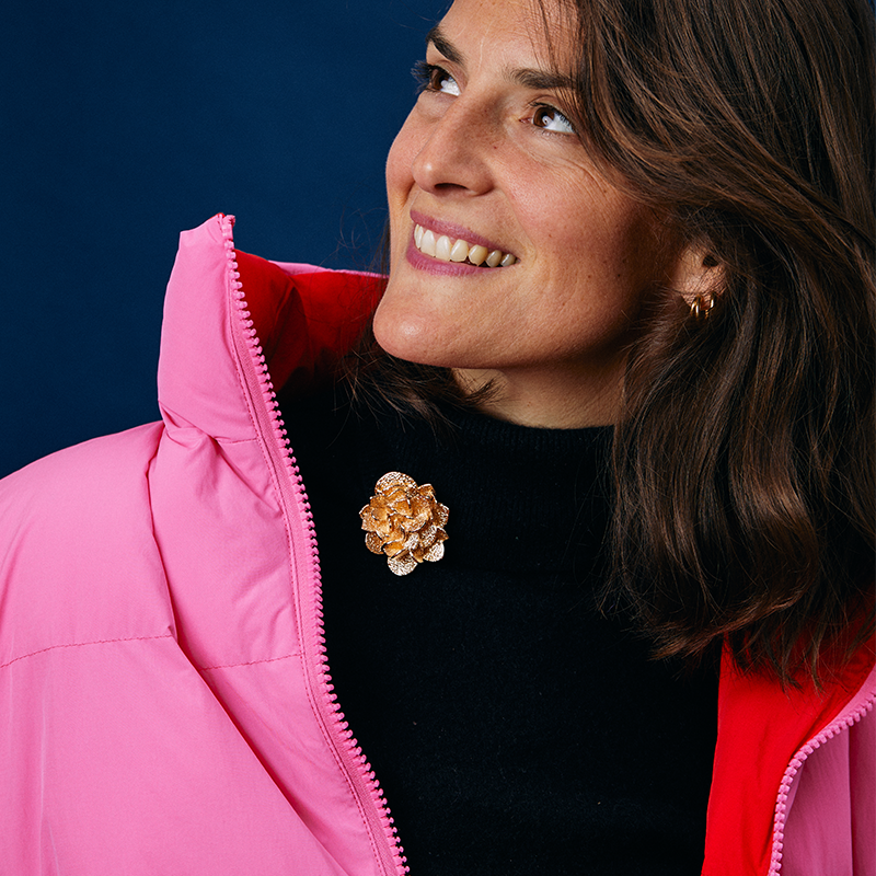 femme souriante portant un clip de couleur doré rose des sables de la marque clap paris. Elle porte une doudoune rose et un col roulé noir